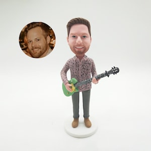 Può includere: Una statuetta bobblehead personalizzata di un uomo che tiene una chitarra verde. La figura indossa una camicia rosa fantasia, pantaloni grigi e scarpe marroni. Una foto di un uomo è in una cornice circolare accanto alla statuetta. La statuetta è su una base bianca.