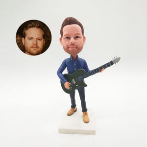 May include: A custom bobblehead figurine of a man playing a black electric guitar. The figure is dressed in a blue shirt, dark jeans, and tan boots. A separate circular image of the person's face is included. The figurine stands on a white base.