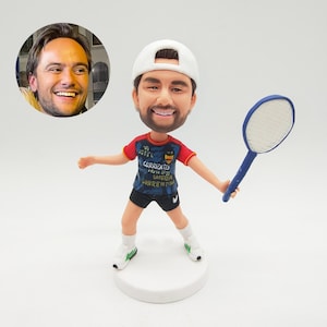 May include: A custom bobblehead figurine of a man in a tennis outfit, holding a blue racket. The figure wears a white cap, a red and blue shirt with text, black shorts, and white trainers. A photo of the man is in the upper left.