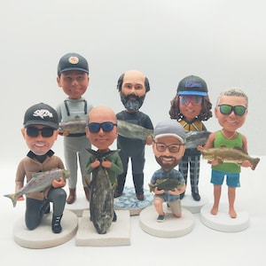 Peut inclure: Plusieurs figurines à tête branlante représentant des hommes tenant des poissons. Les figurines portent des vêtements décontractés, des chapeaux et des lunettes de soleil. Chaque figurine est sur un socle circulaire ou rectangulaire.