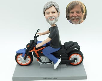 May include: A custom figurine of a person riding a blue and black motorcycle with orange accents. The figurine has a bobblehead of a smiling man with gray hair and a beard. A smaller photo of the man is next to the figurine. The base reads "Road Daddy Steve."