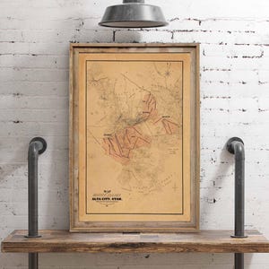 May include: A framed vintage map of Alta City, Utah, displayed on a wooden shelf. The map, with a tan background and red and brown details, is encased in a light brown wooden frame. The text "MAP ALTA CITY, UTAH" is visible at the bottom. The shelf is supported by black pipes.