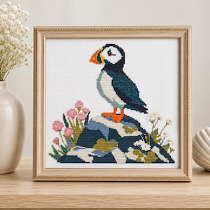 May include: A square framed cross-stitch embroidery featuring a puffin standing on a dark blue rock. The bird has a black and white body with a bright orange beak and feet. Pink and white flowers surround the base of the rock on a white fabric.