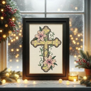 May include: Framed cross-stitch art featuring a cross design with pink lilies and green vines. The cross is gold with a brown border, set against a cream-colored background. The artwork is in a black frame, displayed on a windowsill with festive decorations.
