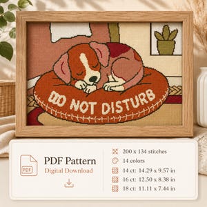Könnte beinhalten: Ein Kreuzstichmuster in einem Holzrahmen zeigt einen braun-weißen Welpen, der auf einem orangefarbenen Kissen mit der Aufschrift DO NOT DISTURB schläft. Darunter listet eine digitale Karte 200 x 134 Stiche, 14 Farben und Maße in cm für 14, 16 und 18 ct.