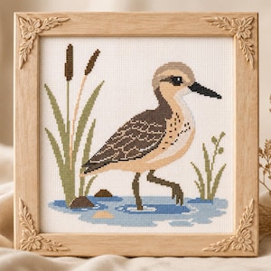 May include: A cross-stitch embroidery of a brown and tan shorebird standing in blue water, surrounded by green cattails and small plants. The artwork is encased in a square, light-colored wooden frame featuring carved floral corner details.