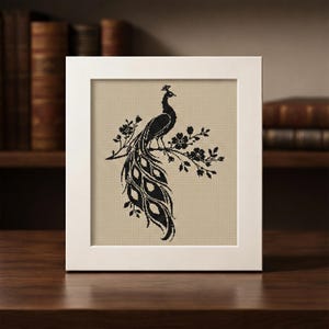May include: A framed cross-stitch artwork featuring a black peacock perched on a flowering branch. The peacock has a long, ornate tail. The artwork is set against a beige background and framed in a white square frame. The artwork is displayed on a dark wooden surface.