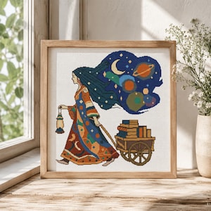 May include: A framed cross-stitch embroidery featuring a person with long dark hair walking while holding a lantern and pulling a wooden cart filled with books. Above the person is a dark blue cloud shape containing a crescent moon and planets.