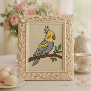 May include: A framed cross-stitch of a cockatiel bird perched on a branch with green leaves and red berries. The bird has yellow, gray, and orange coloring. The frame is cream-colored with a detailed leaf design.