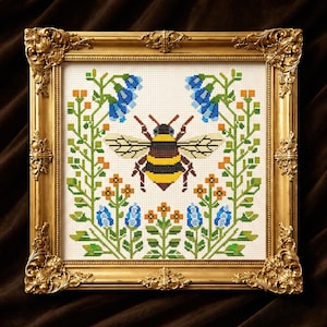 May include: Cross-stitch artwork of a bee with yellow and black stripes, surrounded by blue flowers, orange accents, and green foliage. The framed piece has a gold ornate frame, adding a touch of elegance to the nature-inspired design.
