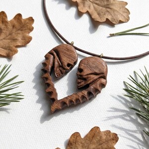 May include: A dark brown wooden pendant carved into a heart shape, featuring two skull profiles facing each other. The pendant is suspended from a thin, dark brown cord, showcasing intricate wood grain textures and detailed skeletal carvings.
