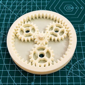 Op de afbeelding: Een crèmekleurige planetaire tandwielset met een grote buitenste ringwiel met interne tanden, drie kleinere planeetwielen en een centraal zonnewiel. De mechanische assemblage is gemaakt van een stevig, mat materiaal met zichtbare tandwieltanden.