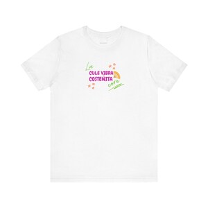 May include: White t-shirt with the text "La Cule Vibra Costenita core" in green, pink, and orange. The design includes a fan graphic and star shapes. The shirt is a casual wear item.