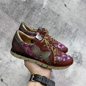 May include: A pair of burgundy sneakers with silver accents. The sneakers feature a star-shaped design on the side, sequin detailing, and tan laces. The soles are beige and the shoes appear to be made of leather.