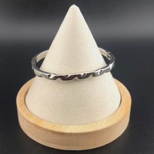 May include: A silver-colored bangle bracelet with a wavy design. The bracelet is displayed on a white cone-shaped stand with a wooden base, set against a dark background. The bracelet's surface reflects light.