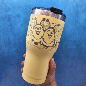 May include: A yellow stainless steel tumbler with a black lid and a design of two corgis dressed as bees. The corgis are wearing bee wings and have black and yellow stripes.