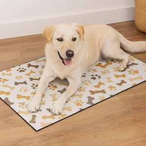 May include: A light-colored Labrador Retriever dog lying on a rectangular pet mat. The mat has a white background with a pattern of brown and gold paw prints and bones. The dog is wearing a black collar.