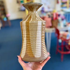 May include: A handcrafted ceramic vase with a flared top and a textured, ribbed body. The vase features vertical stripes in shades of brown and cream. The vase is held in a hand, showcasing its size and design.