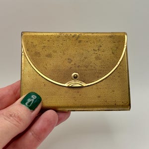 May include: A rectangular gold-tone metal cigarette case featuring a fine cross-hatched texture across the surface. The case has a curved flap closure with a central circular stud fastener and shows signs of age-related patina and wear.