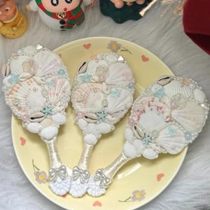 May include: Three decorative hand mirrors with seashell designs. Each mirror features a handle wrapped in white fabric and adorned with a silver bow. The mirror faces are embellished with various seashells, pearls, and small starfish.