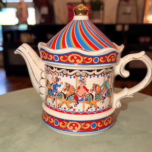 May include: A ceramic teapot featuring a carousel theme with a multi-colored striped lid topped with a gold-colored finial. The body displays illustrations of children riding horses, accented by red, blue, and gold decorative borders and a gold-trimmed handle.