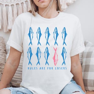 May include: Cream-colored t-shirt with a blue and pink fish graphic. The design features rows of blue fish, with one pink fish standing out. The text "RULES ARE FOR LOSERS" is printed below the fish.