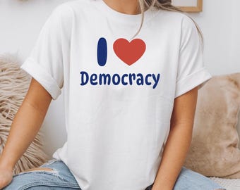 Pode incluir: Camiseta branca com a frase "I ❤️ Democracy" em azul marinho, com uma barra vertical azul e um coração vermelho substituindo a palavra "love". A camisa tem mangas enroladas e é feita de um material macio.