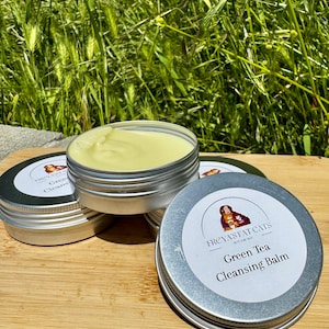 May include: A silver tin with a white label that reads "Freyja's Fat Cats" and "Green Tea Cleansing Balm". The tin is filled with a light green balm.
