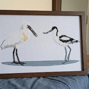 May include: A framed cross-stitch embroidery featuring two birds standing on a blue oval shape. The bird on the left is white with a yellow patch and a long dark beak, while the bird on the right is white with black markings and a long thin beak.