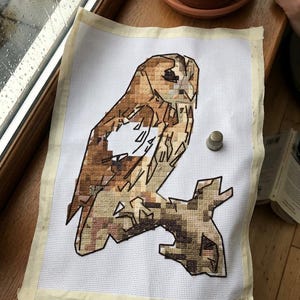 May include: A cross-stitch project on white Aida fabric featuring a geometric owl perched on a branch, rendered in shades of brown, tan, and cream. A small metal thimble sits on the fabric next to the owl, which has a large unstitched white area.