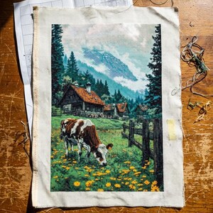 May include: Cross-stitch embroidery of a rural landscape featuring a brown and white cow grazing in a field of yellow flowers. The scene includes a wooden fence, houses, and mountains in the background. The embroidery is on a white fabric.