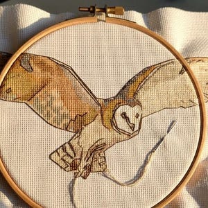 May include: A cross-stitch project featuring a barn owl in flight with outstretched wings, rendered in shades of brown, tan, and white thread on cream-colored Aida fabric. The fabric is secured within a circular wooden embroidery hoop.