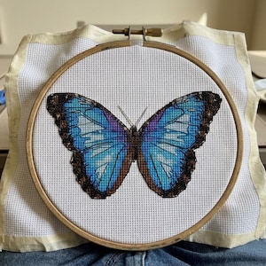 May include: A cross-stitch project featuring a vibrant blue butterfly with dark brown wing edges, centered within a circular wooden embroidery hoop. The design is stitched onto white Aida fabric, with a length of blue embroidery floss resting nearby.