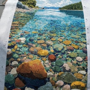 Pode incluir: Bordado em ponto cruz retratando uma serena paisagem aquática. A cena apresenta um leito de rio turquesa claro cheio de pedras coloridas. O céu é azul brilhante com nuvens brancas fofas. As árvores verdes margeiam a costa distante.