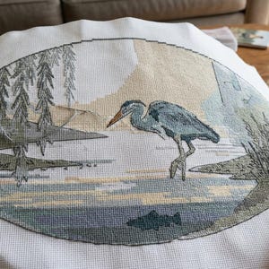 May include: A white fabric piece featuring an oval-shaped cross-stitch embroidery of a blue heron standing in shallow water near a shoreline with trees. A small dark fish is visible beneath the water surface, rendered in shades of blue, green, and tan.