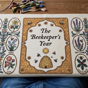 Pode incluir: Um painel de tecido retangular na cor creme com uma borda central de favo de mel dourado com o texto The Beekeeper's Year, uma colmeia e um sol. Doze molduras ovais cercam o centro, cada uma representando uma flor e abelha mensal.