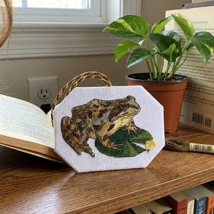 May include: An octagonal white fabric piece featuring a detailed cross-stitch embroidery of a brown and tan frog sitting on a green lily pad. The embroidery is finished with a gold and brown twisted cord hanger attached to the top edge.