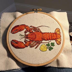 May include: A cross-stitch embroidery project featuring a vibrant orange-red lobster centered within a circular wooden hoop. The design includes a small yellow lemon wedge and a sprig of green parsley positioned below the lobster on light beige fabric.