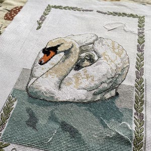 May include: A cross-stitch embroidery project on white fabric featuring a white swan with an orange beak and a small cygnet tucked under its wing. The design is framed by a green leaf border and includes the text Cygnus olor - The Mute Swan.