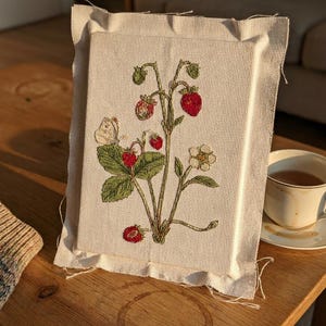 May include: A rectangular piece of off-white Aida fabric featuring a detailed embroidery of a strawberry plant. The design includes green leaves, red strawberries, a small white flower, and a tiny butterfly, all stitched onto the fabric.