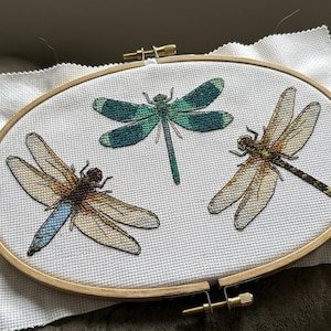May include: An oval wooden embroidery hoop containing white fabric with three embroidered dragonflies. One dragonfly is teal and green, one is blue and tan, and one is brown and tan. A thread organizer and a small ruler are visible nearby.