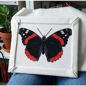 May include: A square piece of off-white Aida fabric featuring a detailed cross-stitch embroidery of an Admiral butterfly. The butterfly has black wings with vibrant red bands and white accents. The fabric is stretched over a square frame.