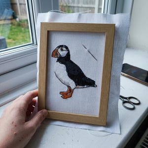 May include: A cross-stitch embroidery of a puffin featuring black, white, and orange thread colors on white Aida fabric. The embroidery is contained within a light-colored wooden frame, and a single silver sewing needle is visible on the fabric.