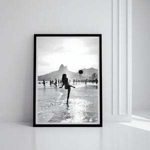 May include: Black and white framed photograph of a beach scene. A person is kicking a ball in the water, with other people in the background. Mountains and buildings are visible in the distance. The image is in a black frame.