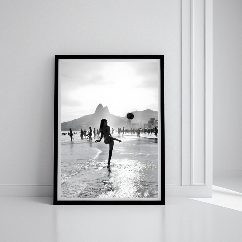 May include: Black and white framed photograph of a beach scene. A person is kicking a ball in the water, with other people in the background. Mountains and buildings are visible in the distance. The image is in a black frame.