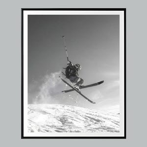 May include: A black and white photograph of a skier mid-air performing a jump above a snow-covered slope. The skier wears a helmet, goggles, and a backpack, with skis crossed in the air and one ski pole held high against a bright sky.