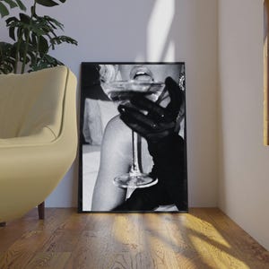 May include: Black and white framed print featuring a person holding a cocktail glass. The person is wearing a black glove. The image is a close-up, with the focus on the glass and the person's mouth.