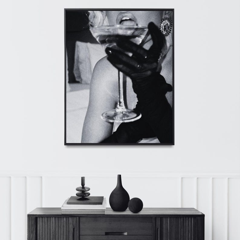 May include: Black and white framed print of a person holding a martini glass. The person is wearing black gloves and an earring. The image is a close-up shot, with the focus on the glass and the person's face.