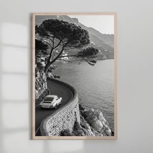 Op de afbeelding: Zwart-wit ingelijste print van een kronkelende kustweg met een witte sportwagen. Een grote boom hangt over de weg, met de zee en bergen op de achtergrond. Het beeld is een schilderachtig uitzicht.
