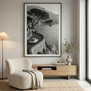 Op de afbeelding: Zwart-wit ingelijste print van een kustweg met een oldtimer. De kronkelende weg volgt de kustlijn, met een stenen muur en de zee. Een boom hangt over de weg. De print wordt boven een houten kast weergegeven.